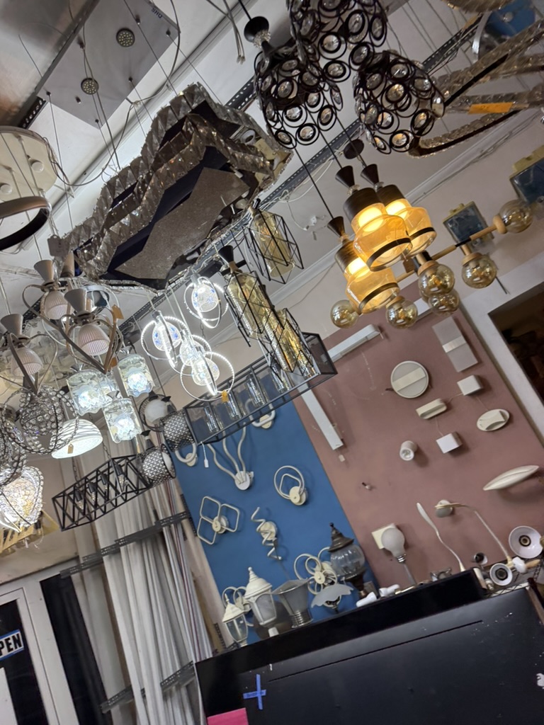 Chandeliers and pendant lamps packed onto ceiling racks, with wall fittings pinned to painted boards behind the counter.